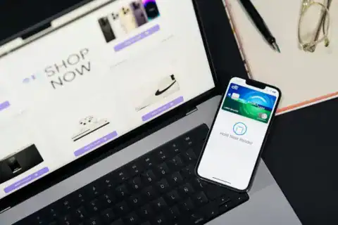 A high-angle shot of a person using a smartphone and laptop to shop online, with glasses and a pen nearby.