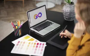  A graphic designer uses a stylus on a drawing tablet, creating a logo on a laptop screen. Color swatches and pencils are on 