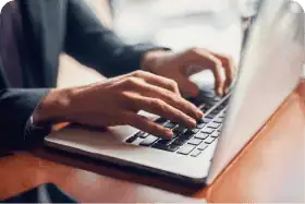 On voit les mains d'une personne qui tape sur le clavier d'un ordinateur portable argenté, avec une tasse de café à proximité