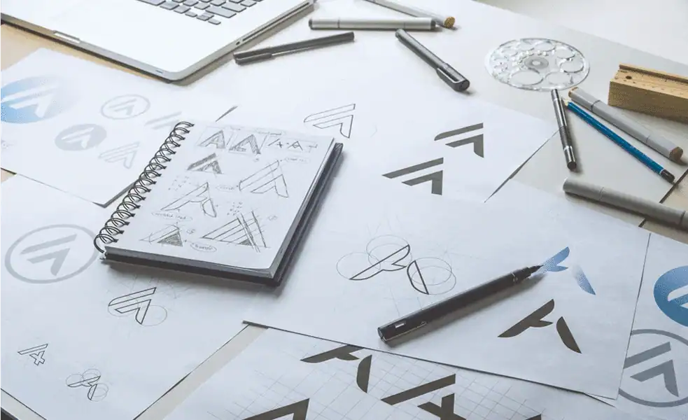 Workspace with sketches and drafts of logo designs on paper, featuring geometric shapes and letter-based concepts.
